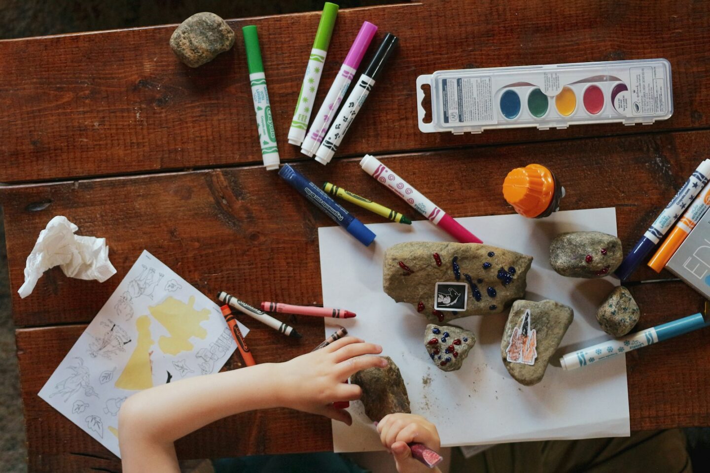 Child painting stones with markers and watercolours on a wooden table (stock photo, not a Shoishob session)
